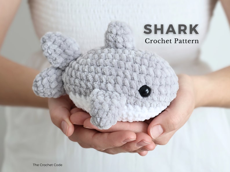 May include: A gray and white crocheted shark toy with black eyes and fins. The text "SHARK Crochet Pattern" is displayed above the shark. The Crochet Code is written at the bottom left.