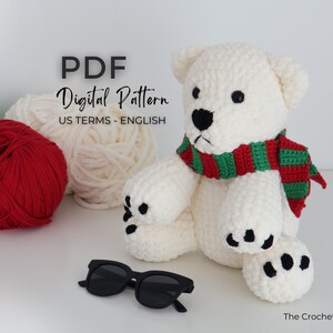 May include: A crocheted polar bear wearing a red and green scarf, with black sunglasses in front. The bear has black eyes and paw pads. Balls of yarn in red, green, and white are in the background. Text on the image reads "PDF Digital Pattern US Terms - English".