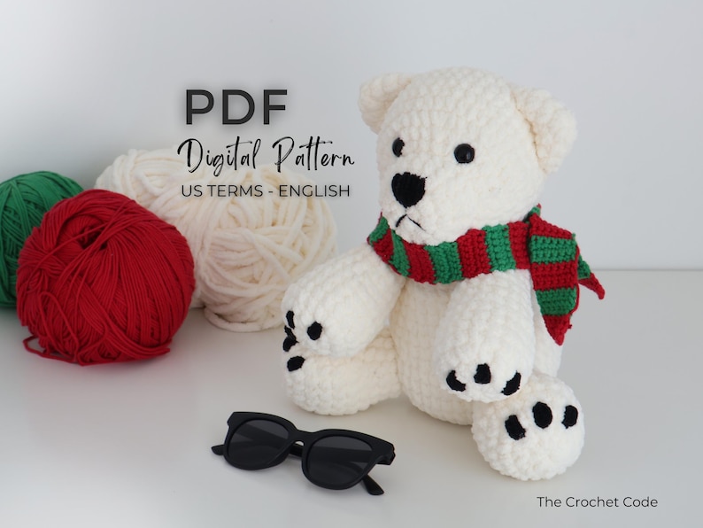 May include: A crocheted polar bear wearing a red and green scarf, with black sunglasses in front. The bear has black eyes and paw pads. Balls of yarn in red, green, and white are in the background. Text on the image reads "PDF Digital Pattern US Terms - English".