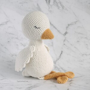 May include: A white crocheted goose with a closed eye, a yellow beak, and orange legs. The goose is sitting on a white marble surface.