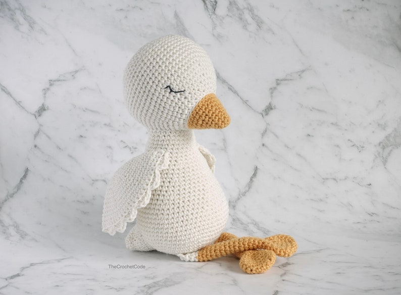 May include: A white crocheted goose with a closed eye, a yellow beak, and orange legs. The goose is sitting on a white marble surface.