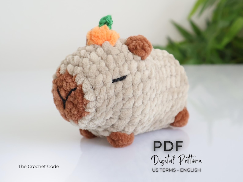 K&ouml;nnte beinhalten: Ein geh&auml;keltes Amigurumi-Spielzeug in Form eines Schweins mit einem kleinen orangefarbenen K&uuml;rbis auf dem Kopf. Das Schwein ist beige mit braunen Akzenten um die Augen und die Nase. Das Spielzeug ist aus weichem, flauschigem Garn gefertigt. Der Text "PDF Digital Pattern US TERMS - ENGLISH" ist am unteren Rand des Bildes sichtbar.