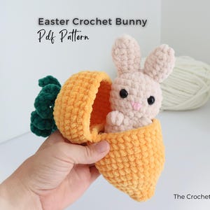 May include: A crocheted Easter bunny peeks out of a plush orange carrot. The bunny is light pink with black eyes and a pink nose. The carrot has green leaves. Text on the image reads "Easter Crochet Bunny Pdf Pattern" and "The Crochet Code".