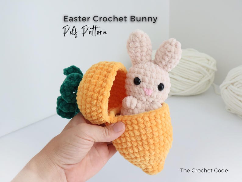 May include: A crocheted Easter bunny peeks out of a plush orange carrot. The bunny is light pink with black eyes and a pink nose. The carrot has green leaves. Text on the image reads "Easter Crochet Bunny Pdf Pattern" and "The Crochet Code".