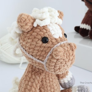 A crocheted horse plushie in shades of brown, white, and gray. The horse has a fluffy mane and tail, black eyes, and a detailed bridle. The plushie is handmade and appears soft and cuddly, perfect for a child's toy or nursery decor.