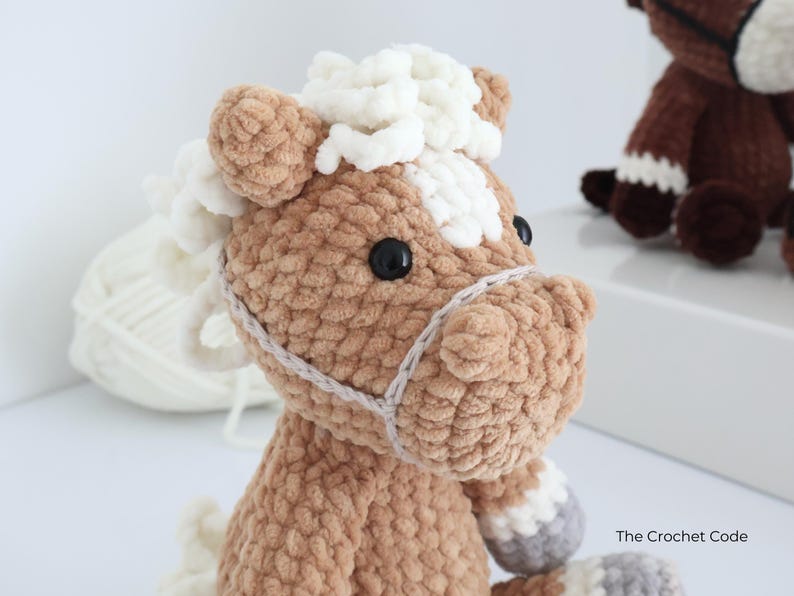 A crocheted horse plushie in shades of brown, white, and gray. The horse has a fluffy mane and tail, black eyes, and a detailed bridle. The plushie is handmade and appears soft and cuddly, perfect for a child's toy or nursery decor.
