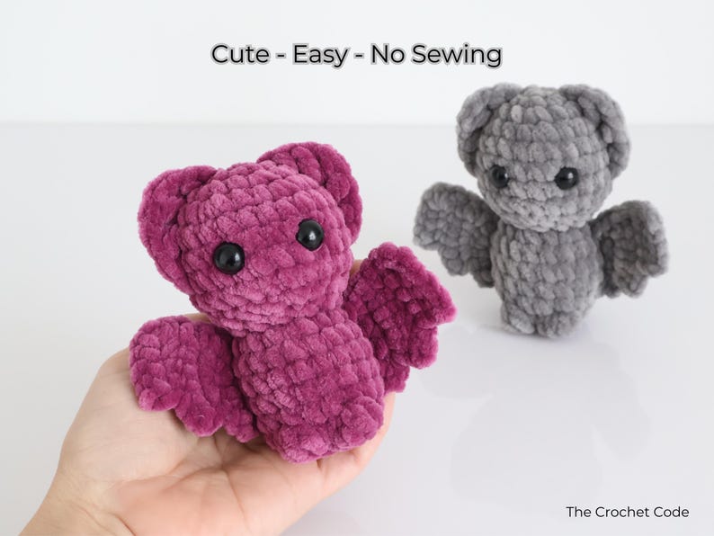 K&ouml;nnte beinhalten: Zwei geh&auml;kelte Fledermaus-Pl&uuml;schtiere. Eines wird in einer Hand gehalten und hat eine tiefmagentafarbene Farbe. Das andere ist grau. Beide haben schwarze Augen und Fl&uuml;gel. Der Text "Cute - Easy - No Sewing" steht oben.