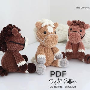 Three plush crochet horse toys in shades of brown, tan, and white. Each horse has a unique mane and facial features, with black bridle details. The image includes the text "The Crochet Code" and "PDF Digital Pattern US TERMS - ENGLISH".
