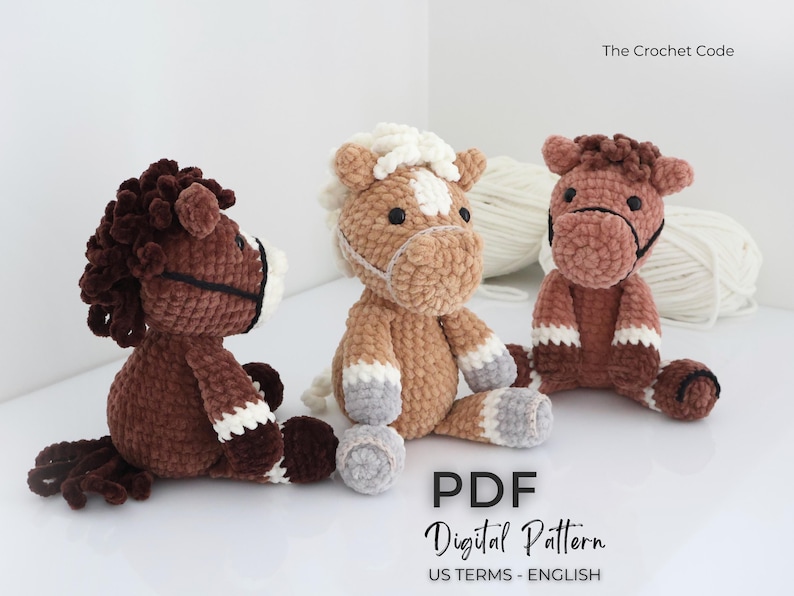 Three plush crochet horse toys in shades of brown, tan, and white. Each horse has a unique mane and facial features, with black bridle details. The image includes the text "The Crochet Code" and "PDF Digital Pattern US TERMS - ENGLISH".