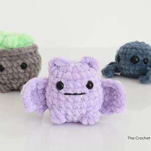 May include: Three handmade crochet plush toys: a grey cauldron with green filling, a lavender bat with wings, and a blue octopus. Each has black button eyes and a smiling mouth. The Crochet Code is in the bottom right.