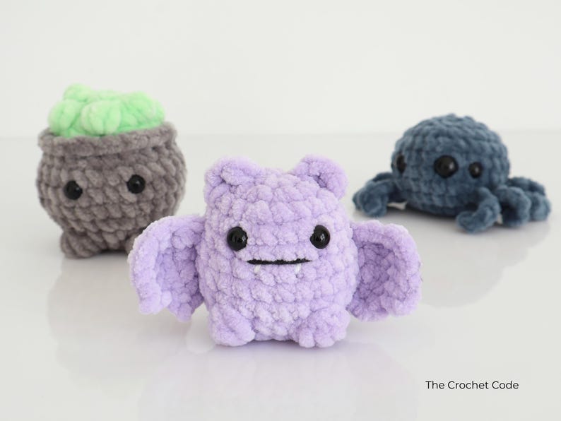 May include: Three handmade crochet plush toys: a grey cauldron with green filling, a lavender bat with wings, and a blue octopus. Each has black button eyes and a smiling mouth. The Crochet Code is in the bottom right.