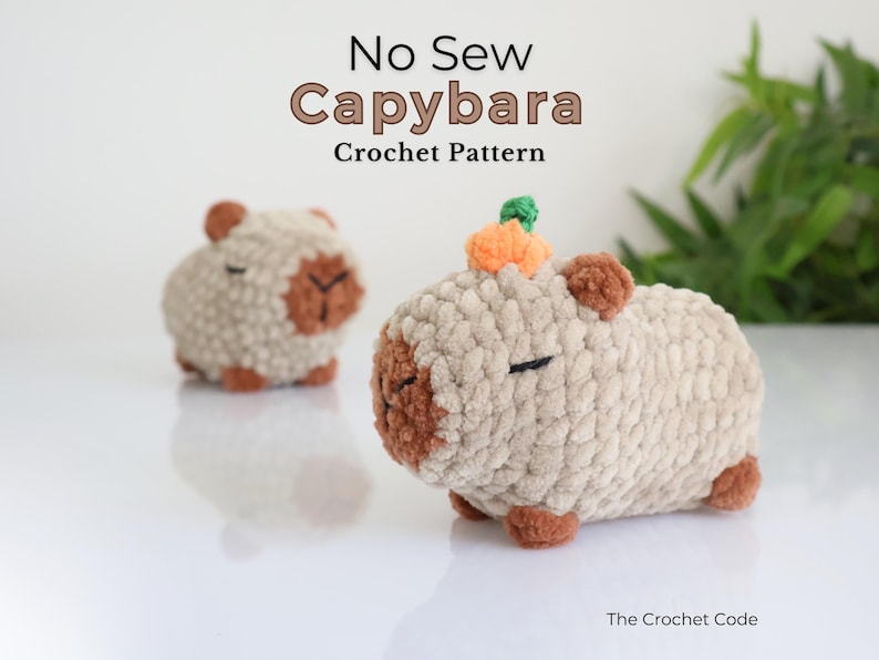 K&ouml;nnte beinhalten: Zwei geh&auml;kelte Capybara-Pl&uuml;schtiere, eines mit einem kleinen orangefarbenen K&uuml;rbis auf dem Kopf. Die Pl&uuml;schtiere sind beige mit braunen Akzenten und stehen auf einer wei&szlig;en Oberfl&auml;che. Der Text "No Sew Capybara Crochet Pattern" befindet sich &uuml;ber den Pl&uuml;schtieren.
