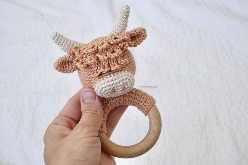 May include: A crocheted cow rattle with a wooden ring. The cow is brown and white with a white face and brown spots. The rattle is ideal for a baby or toddler.