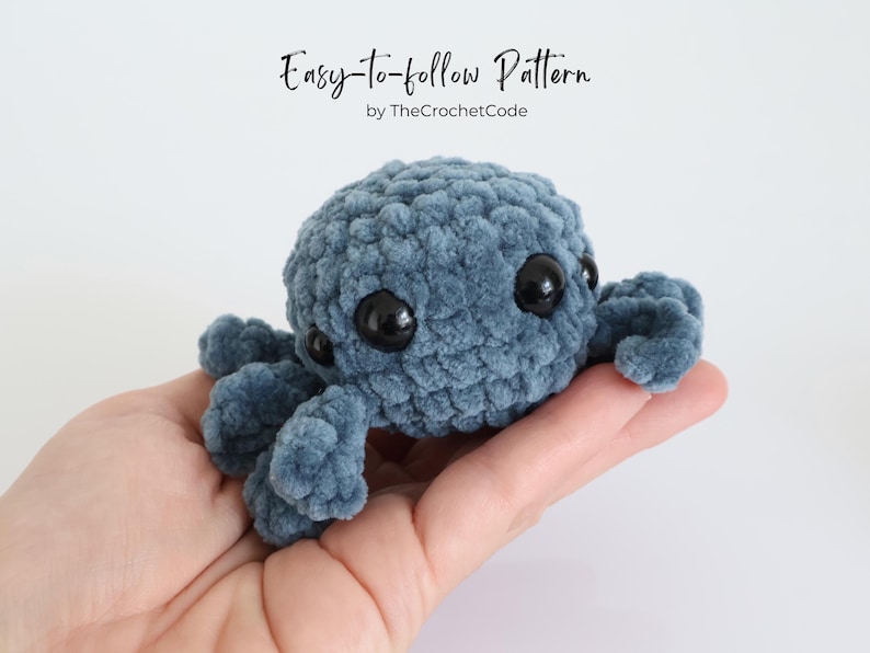 May include: A handmade, blue crocheted octopus with large black eyes and eight tentacles. The text "Easy-to-follow Pattern" and "by TheCrochetCode" are visible in the image. The octopus is held in a person's hand.