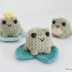 May include: Three handmade crochet frog toys in shades of green, each with black eyes and a smiling mouth. One frog wears a yellow crown. They sit on blue lily pads, perfect for nursery decor.