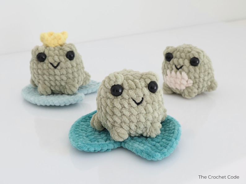May include: Three handmade crochet frog toys in shades of green, each with black eyes and a smiling mouth. One frog wears a yellow crown. They sit on blue lily pads, perfect for nursery decor.