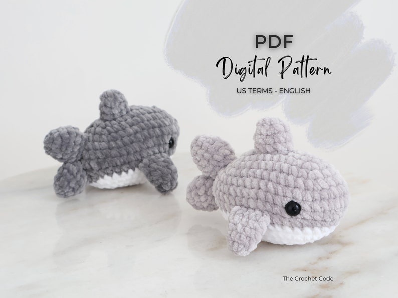 May include: Two plush, gray crocheted shark toys with white bellies and black eyes. The text "PDF Digital Pattern US TERMS - ENGLISH" is displayed above the sharks. The Crochet Code is written at the bottom.