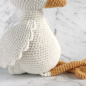 May include: A white crocheted goose with a yellow beak and orange legs. The goose is sitting on a white marble surface.
