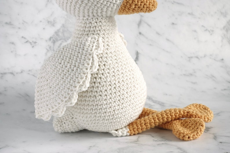 May include: A white crocheted goose with a yellow beak and orange legs. The goose is sitting on a white marble surface.