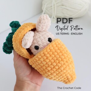 May include: A handmade crochet bunny peeking out of a carrot. The bunny is light pink with black eyes and the carrot is orange with green leaves. The image includes the text "PDF Digital Pattern US Terms - English" and "The Crochet Code".