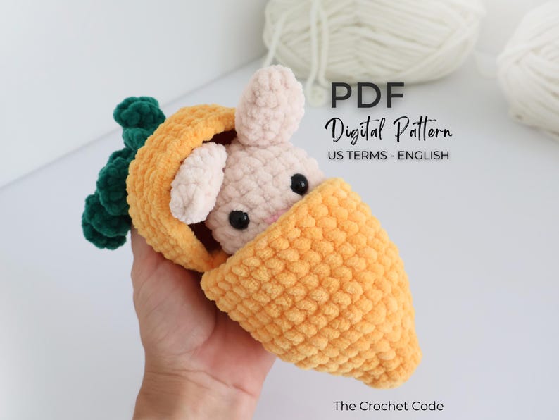 May include: A handmade crochet bunny peeking out of a carrot. The bunny is light pink with black eyes and the carrot is orange with green leaves. The image includes the text "PDF Digital Pattern US Terms - English" and "The Crochet Code".