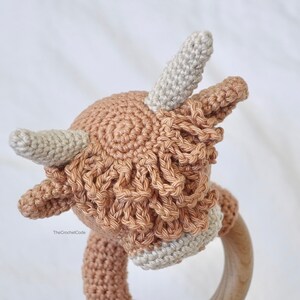 May include: A crocheted brown and white Highland cow rattle toy with a wooden ring. The cow has a fluffy brown mane and white horns.
