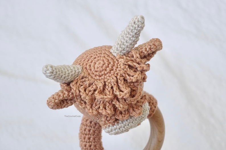 May include: A crocheted brown and white Highland cow rattle toy with a wooden ring. The cow has a fluffy brown mane and white horns.
