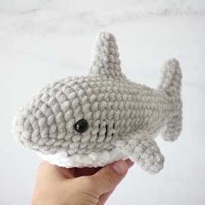 May include: A gray crocheted shark plush toy with white trim around the mouth. The shark has black eyes and a textured surface.