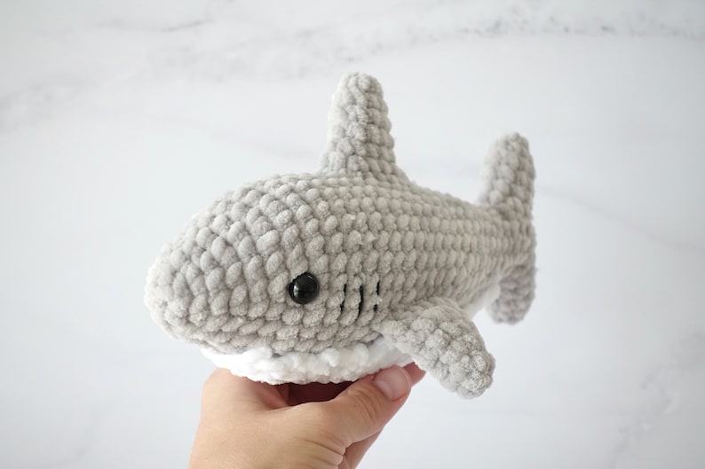 May include: A gray crocheted shark plush toy with white trim around the mouth. The shark has black eyes and a textured surface.