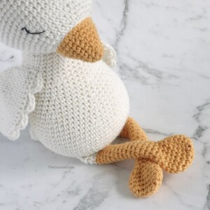May include: A white crocheted amigurumi duck with a yellow beak and feet. The duck is sitting on a white marble surface.
