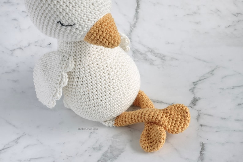 May include: A white crocheted amigurumi duck with a yellow beak and feet. The duck is sitting on a white marble surface.