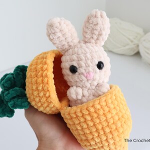 May include: A handmade crochet bunny peeking out of a yellow and orange carrot. The bunny is light beige with black eyes and a pink nose. The carrot has green leaves. The Crochet Code is written on the bottom right.