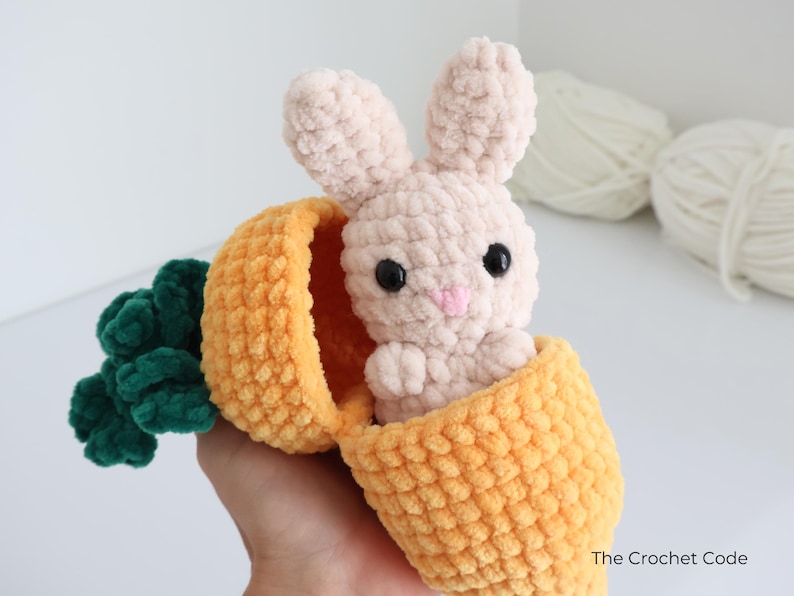 May include: A handmade crochet bunny peeking out of a yellow and orange carrot. The bunny is light beige with black eyes and a pink nose. The carrot has green leaves. The Crochet Code is written on the bottom right.