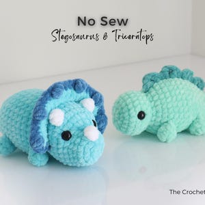 May include: Two crocheted dinosaur toys: a blue Triceratops with white horns and a teal Stegosaurus. The toys are handmade and have black safety eyes. The text "No Sew Stegosaurus & Triceratops" is at the top.