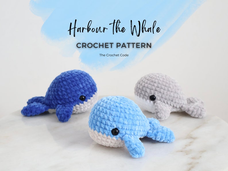 Op de afbeelding: Drie gehaakte walvisknuffels in tinten blauw en grijs, met witte buiken en zwarte ogen. De tekst "Harbour the Whale CROCHET PATTERN" staat boven de walvissen, met "The Crochet Code" eronder.