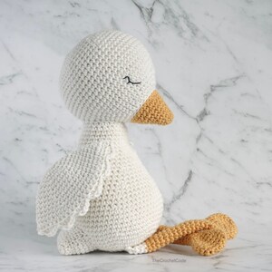 May include: A white crocheted goose with orange feet and beak. The goose is sitting with its head turned to the side and its eyes closed.