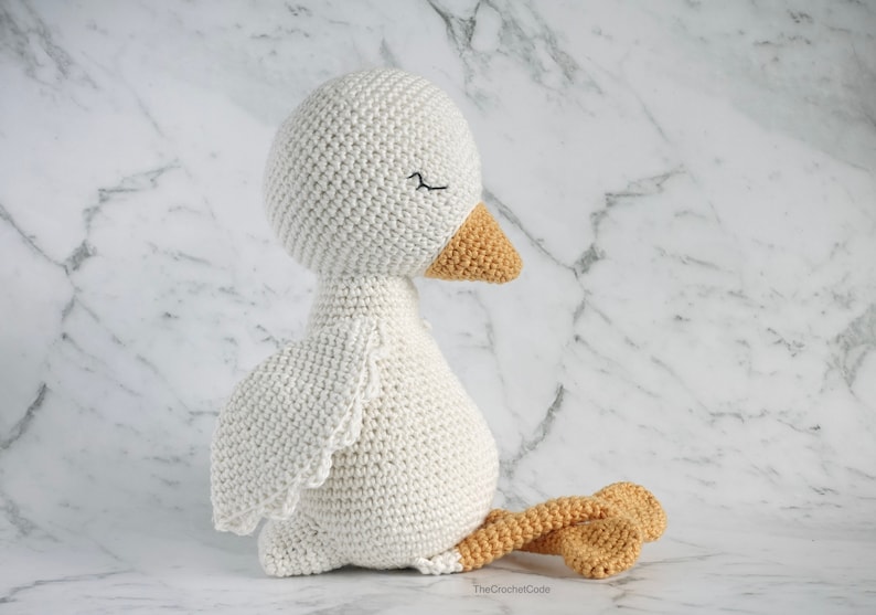 May include: A white crocheted goose with orange feet and beak. The goose is sitting with its head turned to the side and its eyes closed.