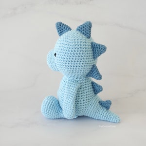 May include: A light blue crocheted dinosaur toy sitting on a white surface. The dinosaur has a long tail and a row of spikes down its back.