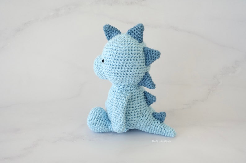May include: A light blue crocheted dinosaur toy sitting on a white surface. The dinosaur has a long tail and a row of spikes down its back.