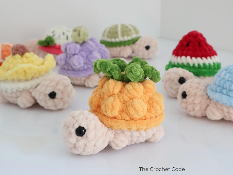 May include: A collection of handmade crochet turtles, each with a unique fruit-themed shell. The turtles feature a variety of colors, including yellow, purple, green, red, and blue. The turtles are made of soft, textured yarn and have black button eyes.