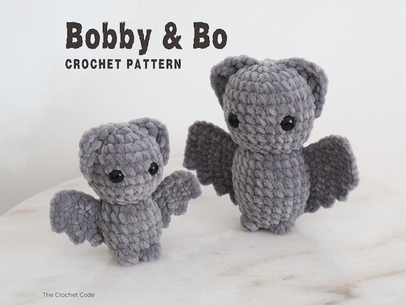 May include: Two gray crocheted bat plushies, one larger than the other, with black eyes. The smaller bat is on the left and the larger bat is on the right. The bats are on a gray surface. The text "Bobby & Bo CROCHET PATTERN" is at the top of the image.