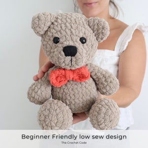May include: A crocheted teddy bear with a brown and gray fur texture, wearing a red bow tie. The bear is being held by a person's hand. Beginner Friendly low sew design. The Crochet Code.