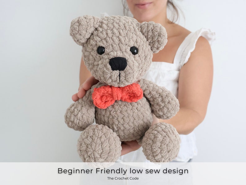 May include: A crocheted teddy bear with a brown and gray fur texture, wearing a red bow tie. The bear is being held by a person's hand. Beginner Friendly low sew design. The Crochet Code.