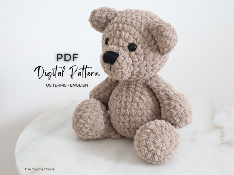 May include: A crocheted teddy bear sitting on a white marble surface. The bear is beige and has black eyes and a black nose. The text "PDF Digital Pattern US TERMS - ENGLISH" is visible in the image.
