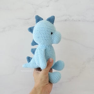 May include: A light blue crocheted dinosaur toy with dark blue accents. The dinosaur has a small black eye and is being held by a hand.