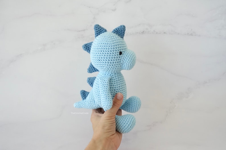 May include: A light blue crocheted dinosaur toy with dark blue accents. The dinosaur has a small black eye and is being held by a hand.