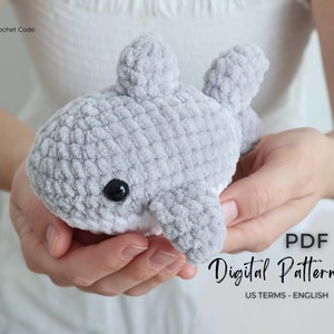 May include: A crocheted gray whale plushie with black eyes and white accents is held in open hands. The whale has fins and a textured surface. Text on the image reads "PDF Digital Pattern US TERMS - ENGLISH".