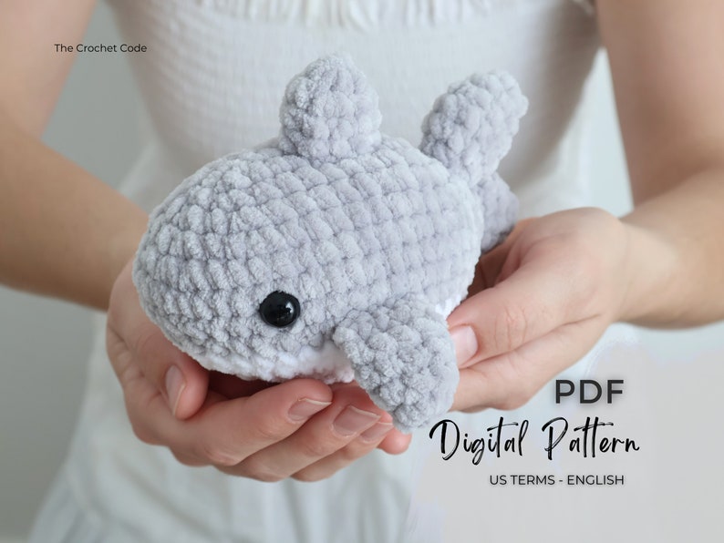 May include: A crocheted gray whale plushie with black eyes and white accents is held in open hands. The whale has fins and a textured surface. Text on the image reads "PDF Digital Pattern US TERMS - ENGLISH".