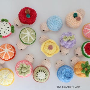 May include: A collection of colorful, crocheted fruit-shaped toys, including a red apple, a green lime, and a pink orange. Each toy has a small, light-colored body. The toys are arranged on a white surface.