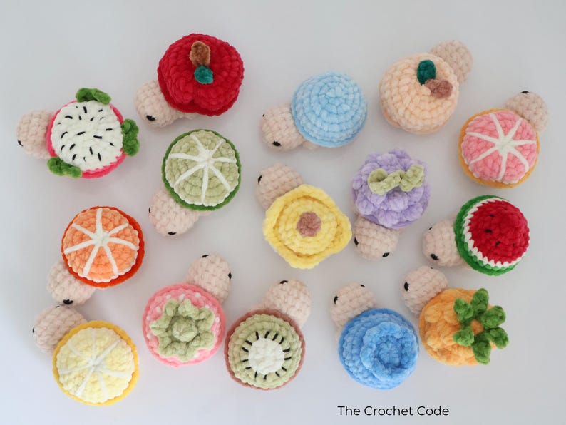May include: A collection of colorful, crocheted fruit-shaped toys, including a red apple, a green lime, and a pink orange. Each toy has a small, light-colored body. The toys are arranged on a white surface.